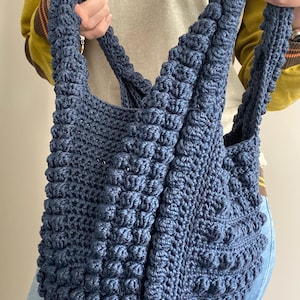 May include: A large, blue crocheted tote bag with a textured, puffy design. The bag has two long straps.