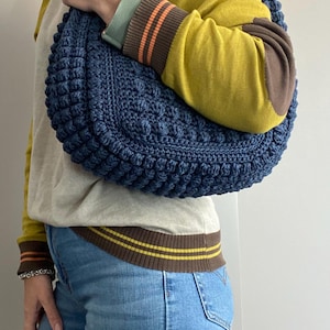 May include: A blue crocheted handbag with a textured, puffy design. The bag is worn over the shoulder of a person wearing a yellow sweater and blue jeans.