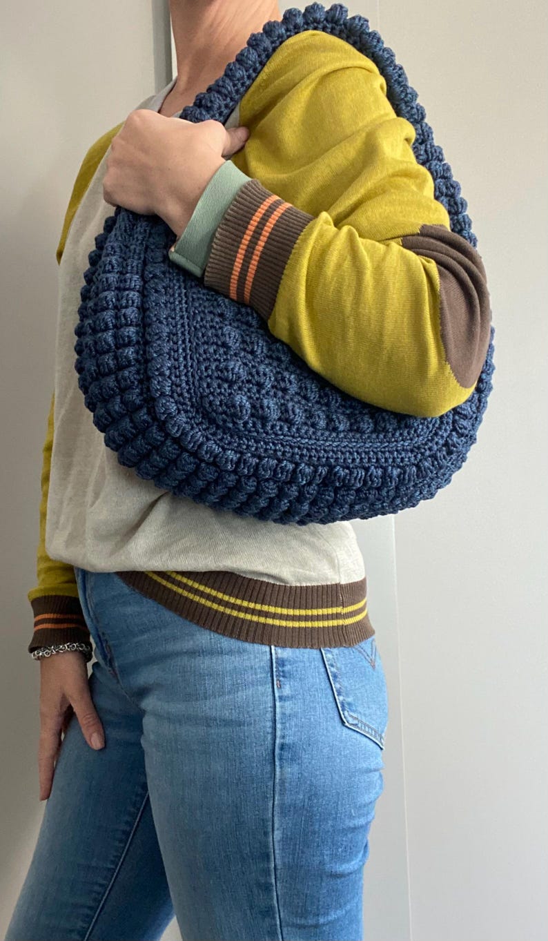 May include: A blue crocheted handbag with a textured, puffy design. The bag is worn over the shoulder of a person wearing a yellow sweater and blue jeans.