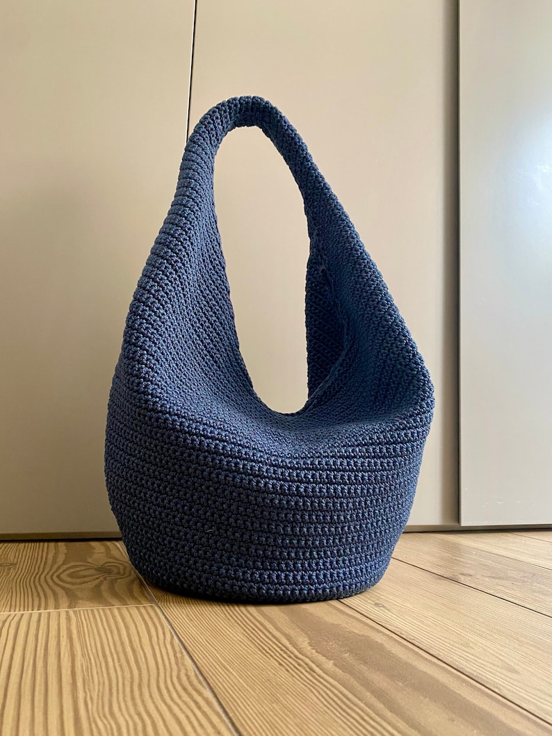 May include: A blue crocheted tote bag with a single strap. The bag is sitting on a wooden floor.