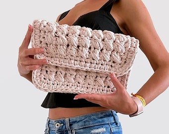 May include: A light pink crocheted clutch bag with a textured design. The bag is held by a person wearing a black top and blue jeans.