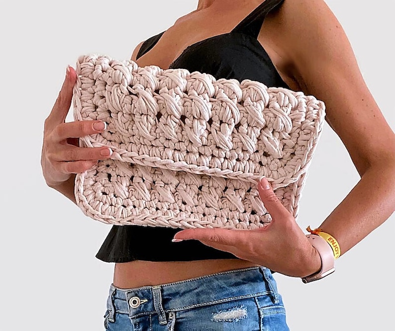 May include: A light pink crocheted clutch bag with a textured design. The bag is held by a person wearing a black top and blue jeans.