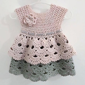 May include: A crocheted baby dress with a pink top and a green bottom. The dress has a ruffled skirt and a flower embellishment on the bodice.