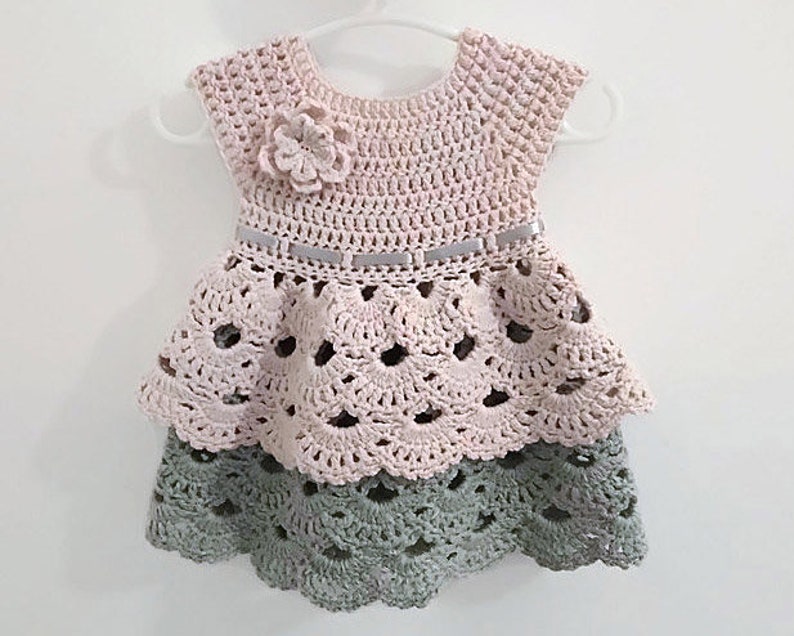 May include: A crocheted baby dress with a pink top and a green bottom. The dress has a ruffled skirt and a flower embellishment on the bodice.