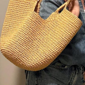 May include: A light tan woven tote bag with two handles. The bag has a rectangular shape and a tight weave, suggesting durability. The bag is being carried over the shoulder.