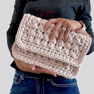 May include: A pale pink crocheted clutch purse with a textured design. The purse is folded in half and held by a person's hands.