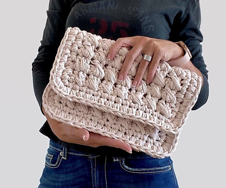 May include: A pale pink crocheted clutch purse with a textured design. The purse is folded in half and held by a person's hands.
