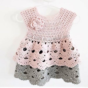 May include: A pink and gray crocheted baby dress with a flower detail and a ribbon around the waist.