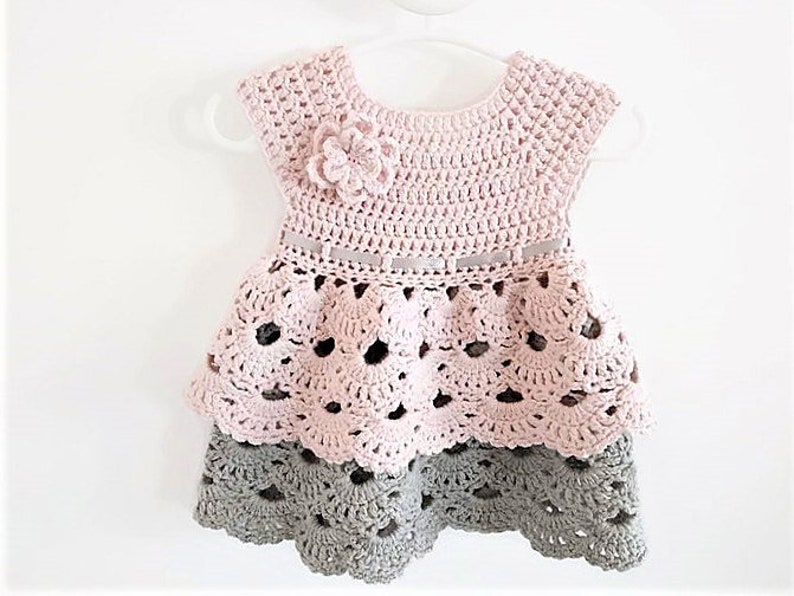 May include: A pink and gray crocheted baby dress with a flower detail and a ribbon around the waist.