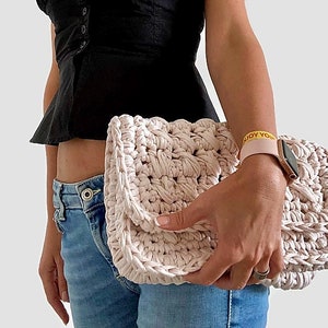 May include: A light pink crocheted clutch bag with a textured design. The bag is held by a person wearing blue jeans and a black top.