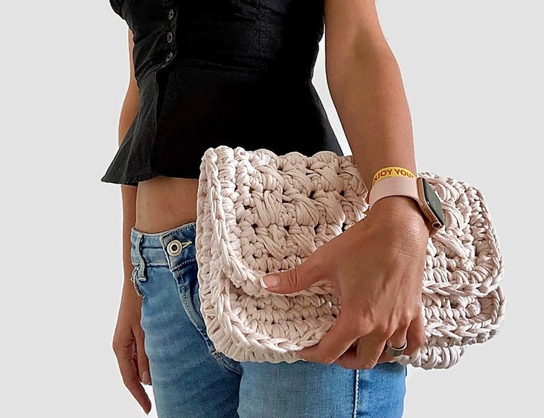 May include: A light pink crocheted clutch bag with a textured design. The bag is held by a person wearing blue jeans and a black top.