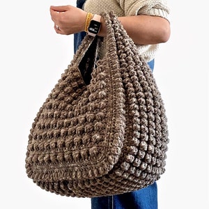 Puede incluir: Un gran bolso de mano de crochet marr&oacute;n con una superficie texturizada y abultada. El bolso tiene dos asas y lo lleva una persona que lleva un jersey y unos vaqueros.