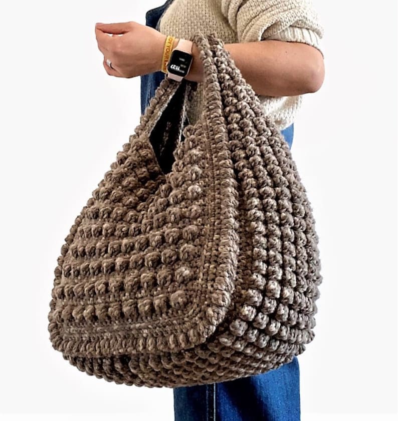 Puede incluir: Un gran bolso de mano de crochet marr&oacute;n con una superficie texturizada y abultada. El bolso tiene dos asas y lo lleva una persona que lleva un jersey y unos vaqueros.