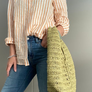 May include: A woman is wearing a striped orange and white linen shirt with blue jeans and a green crocheted tote bag.