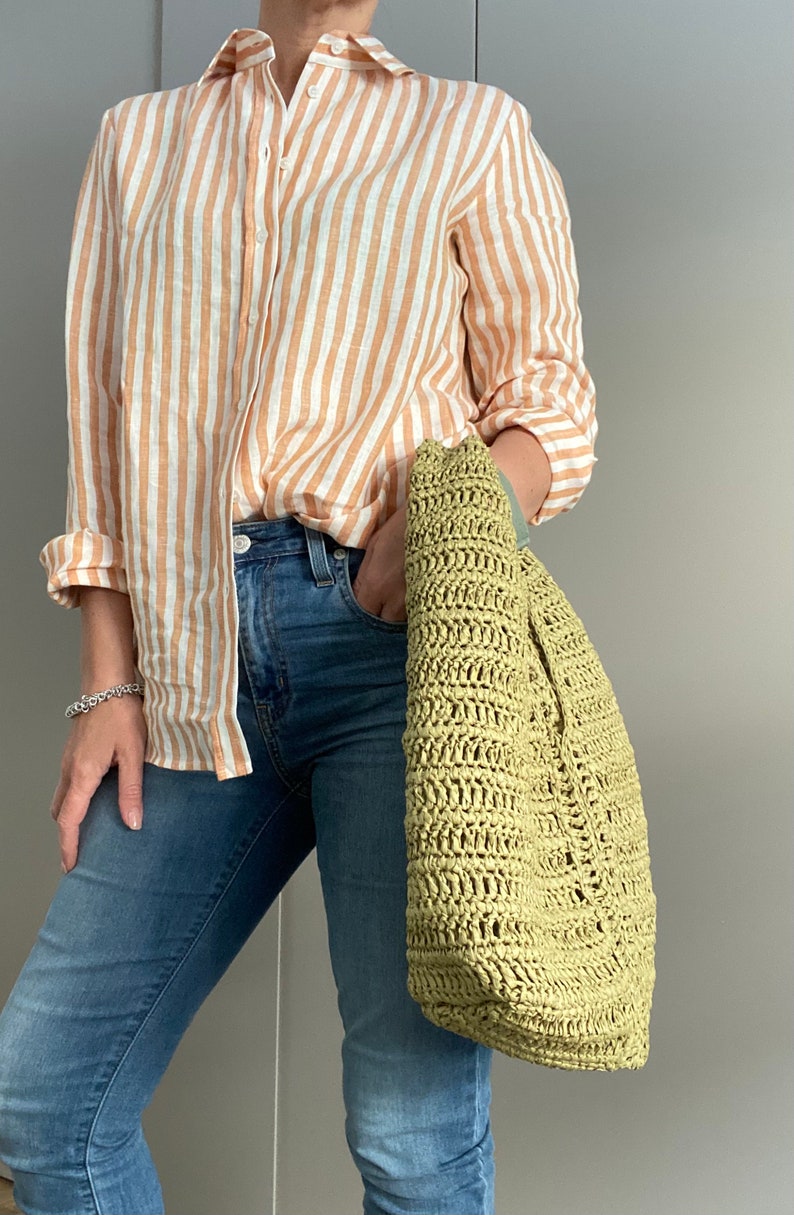 May include: A woman is wearing a striped orange and white linen shirt with blue jeans and a green crocheted tote bag.