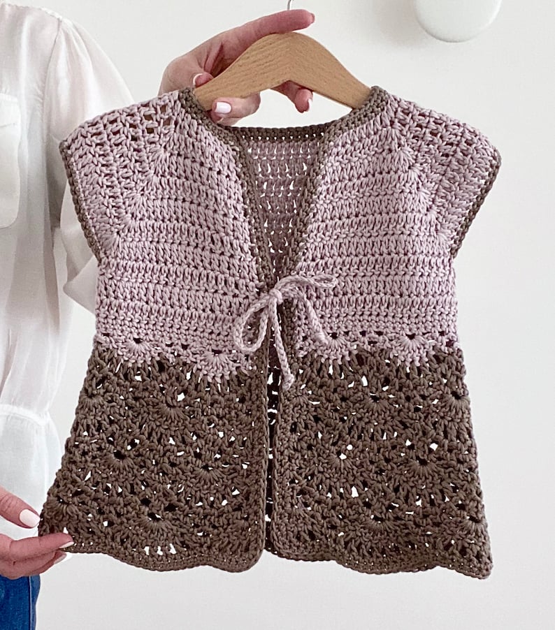 Op de afbeelding: Een gebreid vest met een strik sluiting. Het vest is gemaakt in twee kleuren, licht roze en bruin. Het vest heeft een gegolfde zoom.
