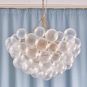 May include: A chandelier featuring numerous clear glass spheres with a swirling ribbed texture, suspended from a central gold-toned metal frame by a matching gold-toned chain. The spherical glass elements are arranged in a dense, clustered formation.