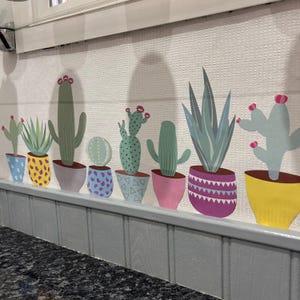 Könnte beinhalten: Eine Reihe von bunten Kakteen und Sukkulenten in Töpfen mit geometrischen Mustern. Die Pflanzen sind an einer weißen Wand mit einem grauen Rahmen angeordnet.