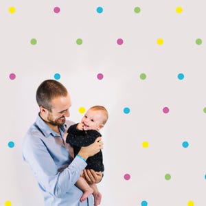 May include: A father holds his baby daughter in his arms. They are both smiling and looking at each other. The background is a white wall with colorful polka dots.