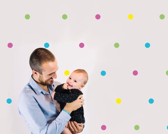 May include: A father holds his baby daughter in his arms. They are both smiling and looking at each other. The background is a white wall with colourful polka dots.