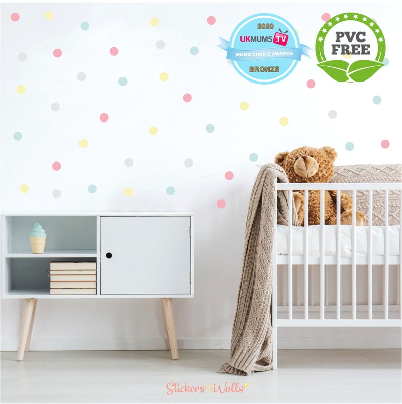 May include: A white wall with pastel colored polka dot wall decals. The decals are in shades of pink, yellow, blue and gray. A white crib with a teddy bear and a blanket is in the background. The text "Stickers 'n' Walls" is at the bottom of the image.