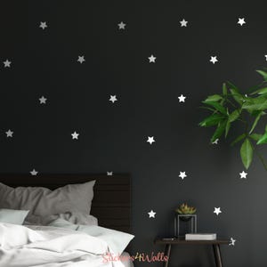 May include: A black wall with white star stickers, a bed with white bedding, a wooden nightstand with a small plant, and the text "Stickers4Walls" on the bottom right corner.