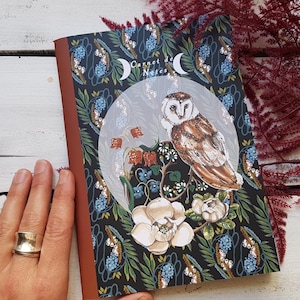 Può includere: Un taccuino con sfondo scuro con un design floreale e lunare, con il testo "Carnet Notes". La copertina presenta un'illustrazione dettagliata di un gufo, fiori e fogliame. Il taccuino ha un dorso marrone ed è posto su una superficie di legno bianco.