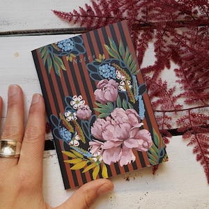 Puede incluir: Un cuaderno floral con peonías rosas y detalles azules, sobre un fondo a rayas marrones. Una mano con un anillo dorado sostiene el cuaderno. Ramas de helechos de color marrón rojizo en el fondo.