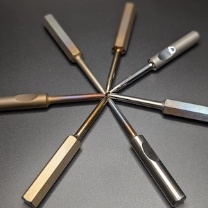 May include: Six metal tools with hexagonal handles and slender shafts are arranged in a starburst pattern against a dark background. The tools have varying finishes, including silver, gold, and iridescent hues, suggesting different materials or treatments.