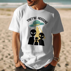 Op de afbeelding: Een wit T-shirt met korte mouwen en een grafische print van drie zwarte buitenaardse silhouetten onder een blauwgroene vliegende schotel. De tekst "THEY'RE HEEEERE" staat in het zwart boven de schotel, die een gele lichtstraal uitstraalt.