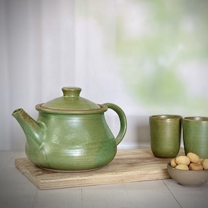 May include: A green ceramic teapot with a lid, two green ceramic mugs, and a bowl of biscuits on a wooden chopping board.