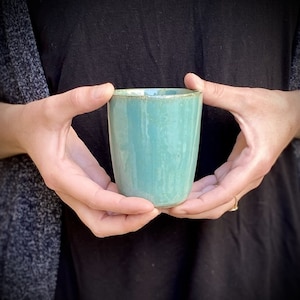 May include: A teal ceramic mug with a light green glaze. The mug is being held by a person's hands.