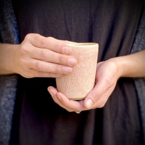May include: A person is holding a small, light pink ceramic cup with a textured surface.