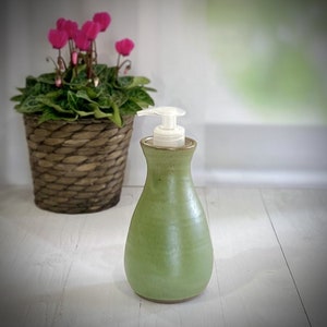 May include: A green ceramic soap dispenser with a white pump. The dispenser has a rounded shape and a light green glaze. A potted plant with pink flowers sits in a woven basket to the left. The background is blurred.