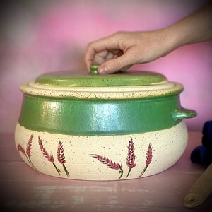 May include: A ceramic cooking pot with a lid. The pot is painted with a green band around the middle and a speckled green lid. The bottom half is off-white with painted purple floral accents. The pot has two small handles.