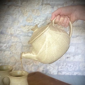 Puede incluir: Una tetera de cerámica beige con tapa se está vertiendo en dos tazas de cerámica beige. La tetera y las tazas están sobre una tabla de cortar de madera.