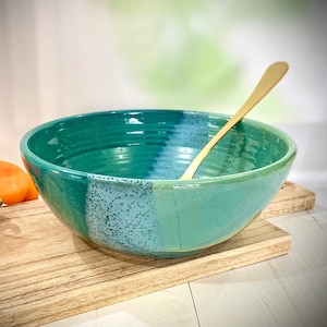 Pode incluir: Uma tigela grande de cerâmica verde-azulada com um esmalte branco e azul. A tigela está sobre uma tábua de cortar de madeira com uma colher de ouro dentro.