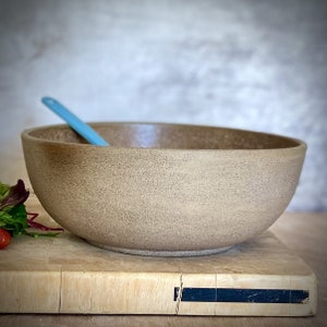 Può includere: Una grande ciotola in ceramica beige macchiata con un cucchiaio azzurro all'interno. La ciotola è appoggiata su un tagliere di legno con una piccola porzione di insalata e un pomodorino visibile. La ciotola ha una forma liscia e arrotondata.