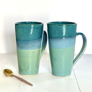 Könnte beinhalten: Zwei Keramikbecher mit einer teal-, blauen und grünen Glasur. Die Becher haben eine abgerundete Form und einen großen Henkel.