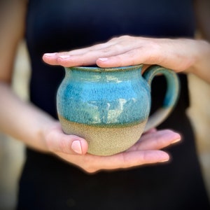 May include: A teal blue and beige ceramic mug with a rounded body and a handle. The mug is glazed with a speckled finish.