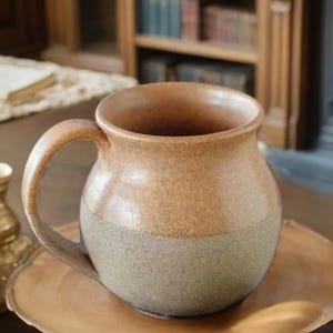 Könnte beinhalten: Eine braune und graue Keramiktasse mit abgerundetem Körper und Henkel. Die Tasse hat eine gesprenkelte Glasur.