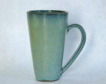 May include: Tall, ceramic mug with a seafoam green glaze and a dark blue rim. The mug has a large handle and a speckled texture. This handmade mug is perfect for coffee, tea, or other beverages.