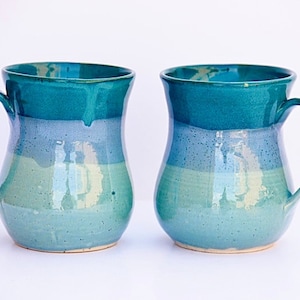 Puede incluir: Dos tazas de cerámica con un esmalte azul claro, verde y blanco. Las tazas tienen un cuerpo redondeado y un asa.