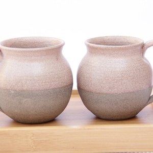 Könnte beinhalten: Zwei Keramikbecher mit hellrosa und grauem Überzug. Die Becher haben einen abgerundeten Körper und einen Henkel.