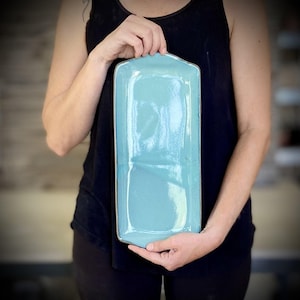 Könnte beinhalten: Ein leicht abgerundetes rechteckiges Serviertablett aus hellblauer Keramik mit einem dunkelgrauen Rand.