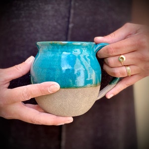 Puede incluir: Una taza de cerámica de color azul verdoso y beige, con forma redondeada y asa. La taza está cubierta con un esmalte brillante.