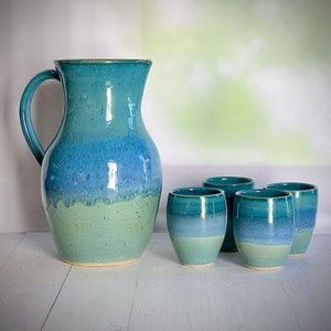 May include: A set of five ceramic pieces in shades of blue and green, including a jug and four cups. The jug has a wide base and a rounded body, while the cups are smaller and more cylindrical. The glaze on all the pieces is uneven, creating a unique and rustic look.