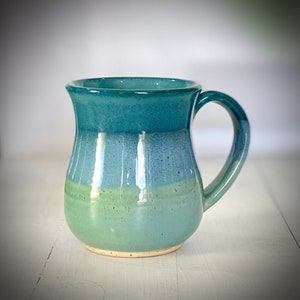 Puede incluir: Una taza de cerámica hecha a mano con un esmalte turquesa, azul y verde. La taza tiene un cuerpo redondeado y un asa.