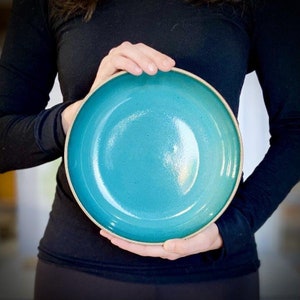 Puede incluir: Un plato de cena grande de cerámica de color turquesa con un borde ligeramente elevado. El plato tiene un acabado liso y brillante.