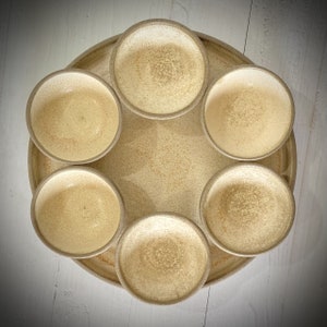 Op de afbeelding: Een set van zes beige keramische kommen, gerangschikt in een cirkel op een beige dienblad. De kommen zijn allemaal even groot en hebben dezelfde vorm, en ze hebben een licht getextureerd oppervlak.